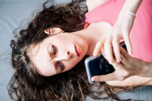 ung kvinna med lång lockigt hår lögner på de soffa och bläddrar social nät på henne telefon med en allvarlig se, topp se foto