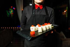 man i svart skjorta och röd rosett slips innehav bricka av sushi foto