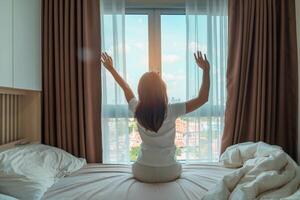 Lycklig kvinna stretching på säng efter vakna upp, ung vuxen kvinna stigande vapen och ser till fönster i de tidigt morgon. färsk koppla av och ha en trevlig dag begrepp foto