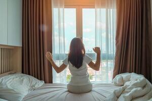 Lycklig kvinna stretching på säng efter vakna upp, ung vuxen kvinna stigande vapen och ser till fönster i de tidigt morgon. färsk koppla av och ha en trevlig dag begrepp foto