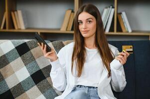 Lycklig ung kvinna innehav telefon och kreditera kort Sammanträde på de soffa. uppkopplad handla. foto