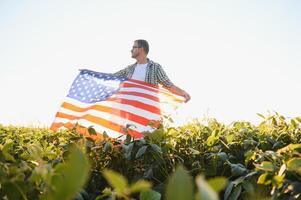 en ung jordbrukare står med en USA flagga i en sojaböna fält. de begrepp av de oss jordbruks industri. foto