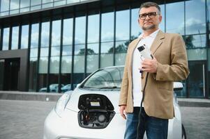 stilig man innehav laddning kabel- på elektrisk laddning station punkt stående nära hans ny bil. foto