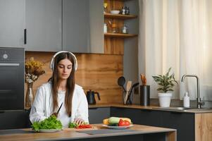 förtjusande trevlig charmig glad Söt skön eleganta upphetsad flicka lyssnande till musik, sång i modern ljus vit kök, matlagning hemlagad maträtt med inhemsk Produkter foto