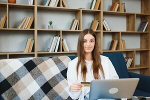 leende kvinna innehav kreditera kort och använder sig av bärbar dator på soffa i levande rum. foto