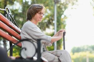 elegant äldre kvinna i de skjorta är Sammanträde på de bänk i en parkera på en värma dag foto