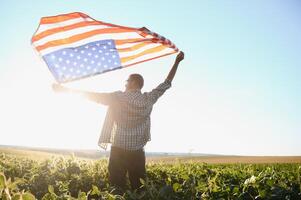 en ung jordbrukare står med en USA flagga i en sojaböna fält. de begrepp av de oss jordbruks industri. foto