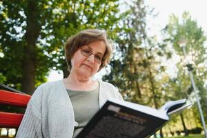porträtt av senior kvinna Sammanträde på en parkera bänk läsning en bok foto