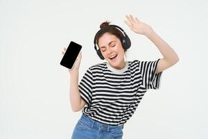 porträtt av eleganta ung kvinna dans i trådlös hörlurar, använder sig av hörlurar och mobil telefon musik app till lyssna till favorit sånger, Framställ över vit bakgrund foto