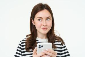 porträtt av flicka med telefon, kvinna innehav smartphone och tänkande, leende och ser åt sidan, beslutar till köpa smth uppkopplad, vit bakgrund foto