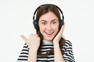 porträtt av upphetsad kvinna i trådlös hörlurar, som visar grymt bra Nyheter, pekande vänster, stående över vit bakgrund foto