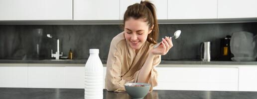 porträtt av Lycklig ung kvinna lutar på kök bänkskiva och äter spannmål, har mjölk och skål i främre av henne, har henne frukost, bär morgonrock foto