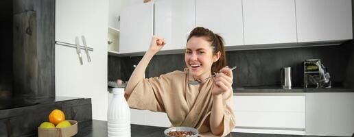 porträtt av Lycklig, skrattande ung kvinna äter spannmål med mjölk, segrar, har frukost och känsla upphetsad, energisk morgon- begrepp foto