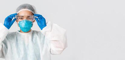 covid19, coronavirus sjukdom, sjukvård arbetare begrepp. seriöst utseende asiatisk kvinna läkare i personlig skyddande Utrustning, sätta på glas innan som förs in i labb, vit bakgrund foto