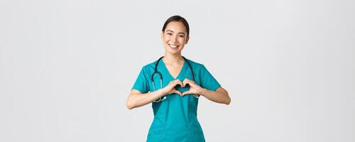 covid19, sjukvård arbetare, pandemi begrepp. härlig omtänksam asiatisk läkare, kvinna sjuksköterska i scrubs som visar hjärta gest och leende, tar vård av patienter med kärlek, vit bakgrund foto