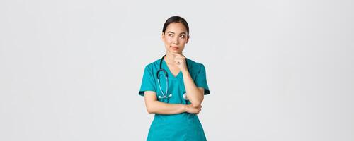 covid19, sjukvård arbetare, pandemi begrepp. omtänksam smart asiatisk sjuksköterska i scrubs ser bort och tänkande, leende nöjd. läkare ha intressant aning, funderar över vit bakgrund foto