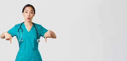 covid19, sjukvård arbetare, pandemi begrepp. förvånad och imponerad asiatisk kvinna sjuksköterska, läkare i scrubs pekande fingrar ner och ser frågade, intresserad i ny kampanj, vit bakgrund foto