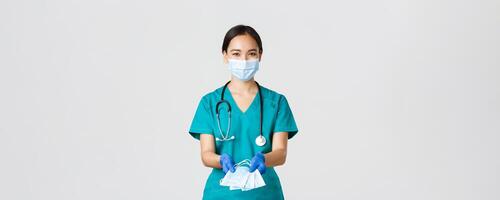 covid19, coronavirus sjukdom, sjukvård arbetare begrepp. vänlig leende asiatisk läkare, kvinna läkare i scrubs och sudd handskar lämnandet över medicinsk masker till patienter, vit bakgrund foto