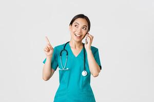 covid19, sjukvård arbetare och förebyggande virus begrepp. porträtt av ung Söt asiatisk kvinna läkare, sjuksköterska i scrubs framställning uppkopplad ordning, talande på telefon och pekande övre vänster hörn, leende foto