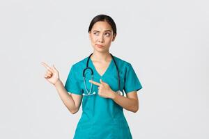 covid19, sjukvård arbetare, pandemi begrepp. skeptisk och tveksam Söt asiatisk läkare, sjuksköterska i scrubs flin, pekande övre vänster hörn, ser med motvillig tveksam ansikte, vit bakgrund foto