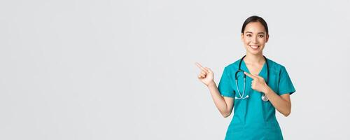 covid19, sjukvård arbetare, pandemi begrepp. leende Söt asiatisk läkare, sjuksköterska i scrubs och stetoskop, pekande fingrar övre vänster hörn, som visar uppkopplad klinik baner, vit bakgrund foto