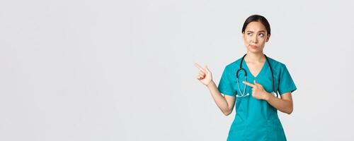 covid19, sjukvård arbetare, pandemi begrepp. skeptisk och tveksam Söt asiatisk läkare, sjuksköterska i scrubs flin, pekande övre vänster hörn, ser med motvillig tveksam ansikte, vit bakgrund foto