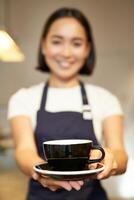 porträtt av leende asiatisk flicka Barista, servitris i Kafé enhetlig, ger du kopp av kaffe, förbereda dryck för klient, ser vänlig foto