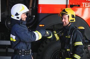 två brandmän i skyddande Kläder i hjälmar med brand motor, vänlig handslag foto