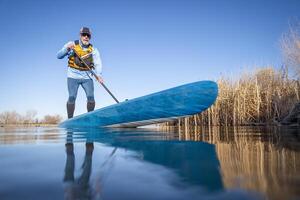 senior manlig paddlare är paddling en stå upp paddelbräda på en lugna sjö i vår, groda perspektiv från ett verkan kamera på vatten nivå foto