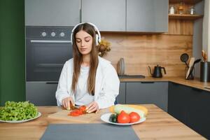 förtjusande trevlig charmig glad Söt skön eleganta upphetsad flicka lyssnande till musik, sång i modern ljus vit kök, matlagning hemlagad maträtt med inhemsk Produkter foto