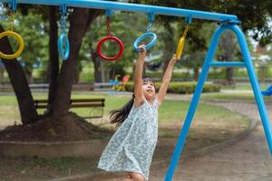Lycklig flicka hängande på apa bar förbi hand håller på med träning. liten asiatisk flicka spelar på utomhus- lekplats i de parkera på sommar semester. friska aktivitet. foto
