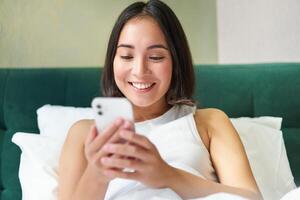 porträtt av leende asiatisk kvinna liggande i säng, ser på smartphone, använder sig av mobil telefon med Lycklig ansikte, avkopplande på lat morgon- foto