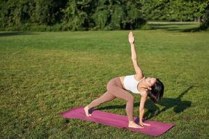 porträtt av ung asiatisk kvinna stretching, håller på med yoga på sudd matta, utövar i parkera, uppmärksam Träning på färsk luft foto