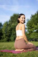 porträtt av asiatisk kvinna Sammanträde och mediterar i parkera, håller på med yoga på färsk luft, kondition övningar, leende på kamera foto