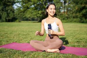 leende kondition flicka sitter på matta och drycker vatten efter träna, Avsluta yoga Träning i parkera, övning på färsk luft foto