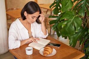 skön ung asiatisk kvinna Sammanträde i Kafé med en bok, äter croissant och läsning, dricka kopp av kaffe foto