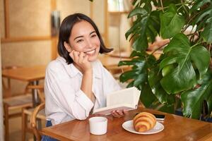 romantisk asiatisk kvinna Sammanträde med bok i Kafé, äter croissant och dricka kaffe, läsning och leende, njuter ensam tid foto