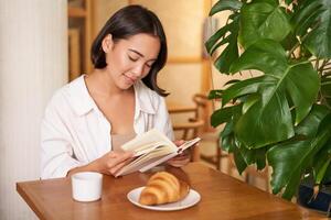 leende ung asiatisk kvinna åtnjuter läsning bok medan sitter ensam i Kafé, drycker kaffe, ser på sida foto