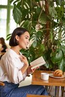 vertikal skott av asiatisk brunett flicka Sammanträde elegant med bok, läsning i Kafé och leende, dricka kaffe foto