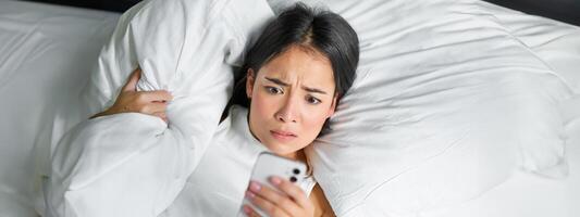 porträtt av asiatisk flicka vaknar upp i morgon, utseende på mobil telefon med chockade ansikte, försov sig, kramas kudde och grimaserande foto