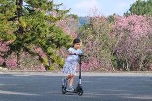 söt liten flicka ridning skoter på gata i utomhus- parkera på sommar dag. Lycklig asiatisk flicka ridning en sparka skoter i de parkera. aktiva fritid och utomhus- sport för barn. foto