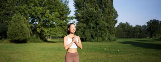 vertikal skott av asiatisk kvinna stående i asana, håller på med yoga övningar på färsk luft i parkera, bär damasker, stående på sudd matta foto