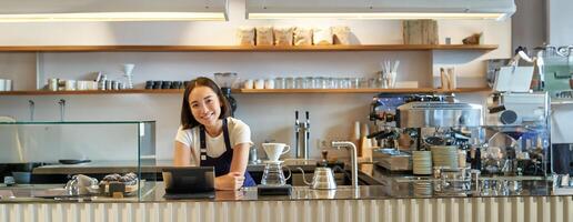 Lycklig leende asiatisk Barista, flicka Bakom disken, arbetssätt med pos terminal och bryggning filtrera utrustning, framställning kaffe i Kafé foto