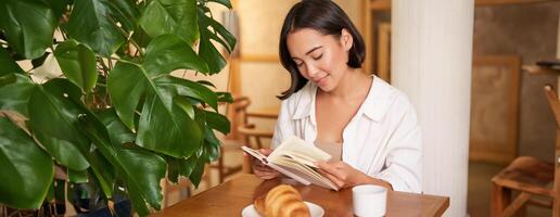 leende ung asiatisk kvinna åtnjuter läsning bok medan sitter ensam i Kafé, drycker kaffe, ser på sida foto