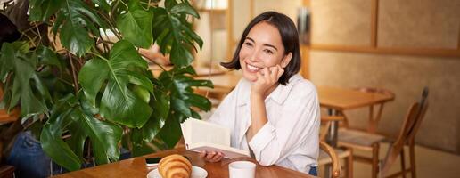 romantisk asiatisk kvinna Sammanträde med bok i Kafé, äter croissant och dricka kaffe, läsning och leende, njuter ensam tid foto