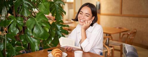 romantisk asiatisk kvinna Sammanträde med bok i Kafé, äter croissant och dricka kaffe, läsning och leende, njuter ensam tid foto