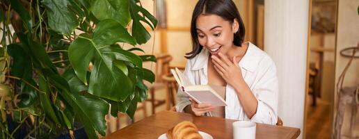 asiatisk flicka utseende förvånad på sidor, läsning med spänning, Sammanträde i Kafé och äter croissant foto