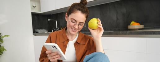 porträtt av eleganta ung kvinna i glasögon, Sammanträde i de kök med mobil telefon och äter äpple, kontroll social media, använder sig av smartphone app foto
