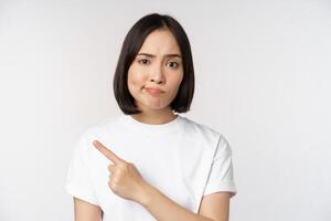 skeptisk asiatisk flicka i vit t-shirt, pekande på produkt eller logotyp med besviken grimas, motvilja och klaga på smth, stående över vit bakgrund foto