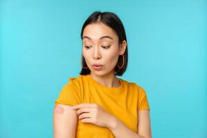 vaccin kampanj från covid19. ung skön, friska asiatisk kvinna som visar axel med plåster, begrepp av vaccination, stående över blå bakgrund foto